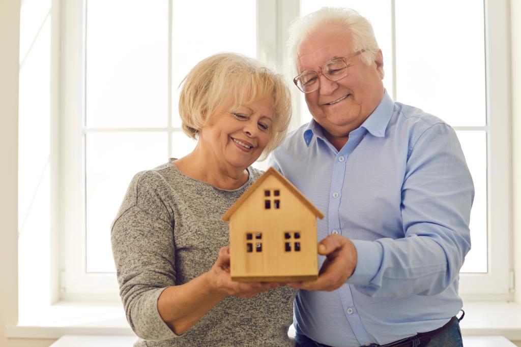 An elderly couple side by side, holding a model of a house. | Featured image for Transition to Retirement Explained - Benefits & Disadvantages blog.